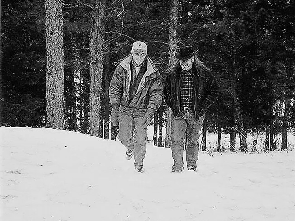 Two men walking through snow among pine trees